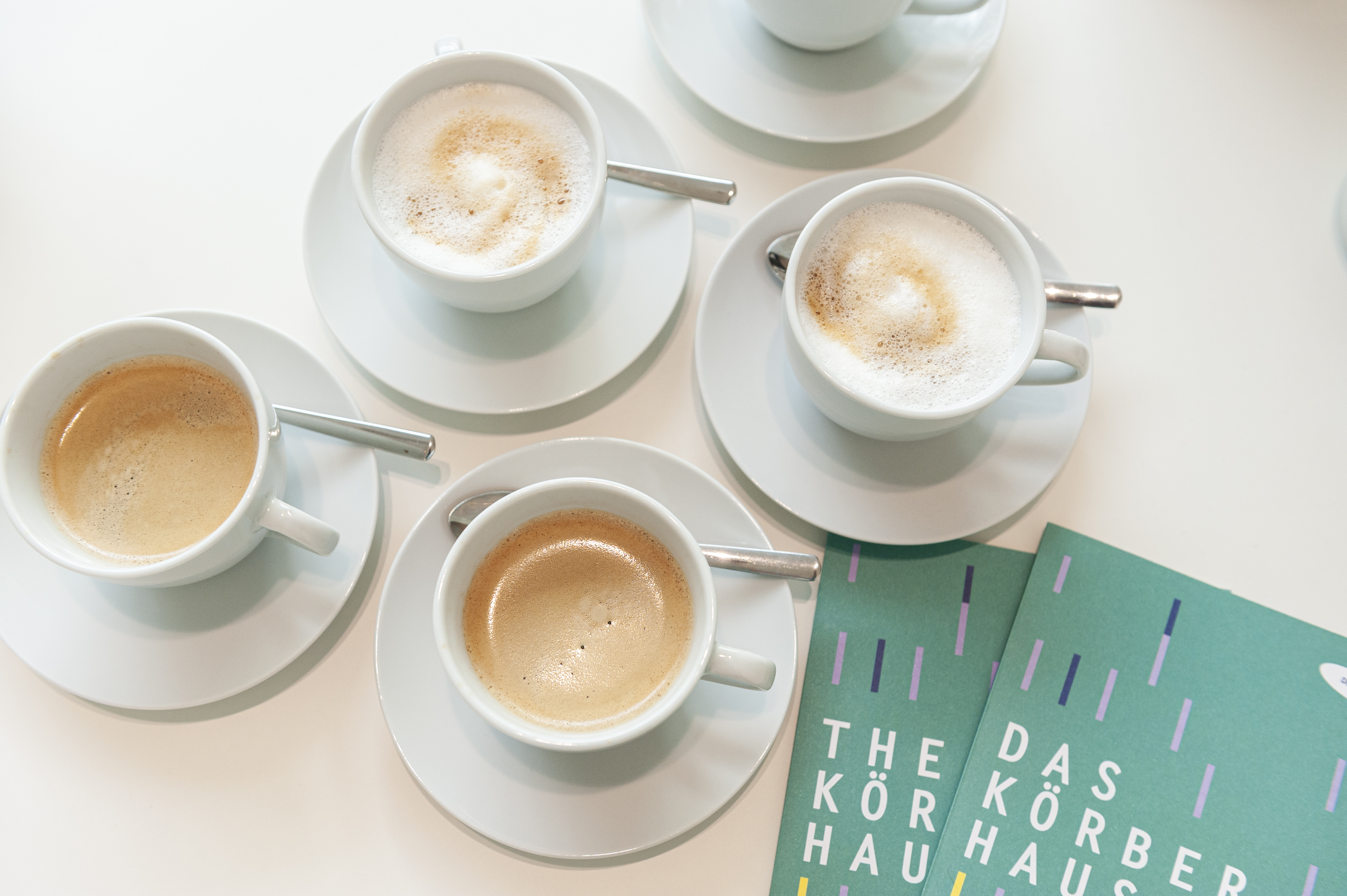 Vier volle Tassen Kaffee von oben fotografiert. Daneben zwei Flyer des KörberHauses.