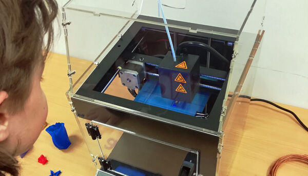 Auf dem Foto ist der 3D-Drucker in seinem Plexiglas-Kasten abgebildet mit einem Kind davor.